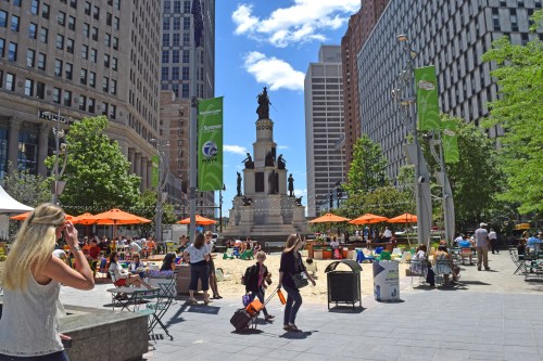 A bustling city square with a monument, people walking, orange umbrellas, and tall buildings under a clear blue sky.
