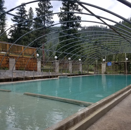 A covered swimming pool surrounded by trees and mountains, with a clear blue sky above.