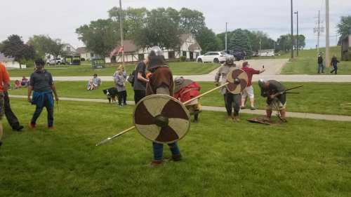 A group of people in historical costumes with shields and spears, participating in a reenactment on a grassy area.