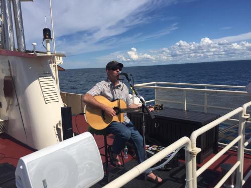A musician plays guitar and sings on a boat deck, with a scenic ocean view and blue sky in the background.