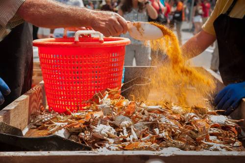 Hands pouring seasoning over a basket of crabs at a seafood festival, with people in the background.