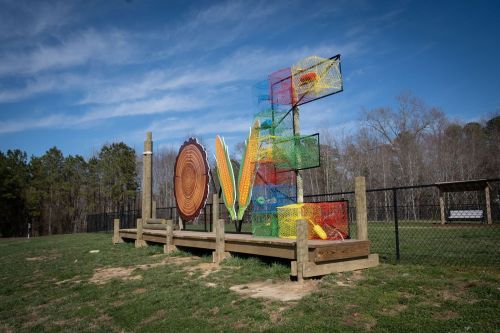 Colorful climbing structure with nets and wooden elements in a grassy area, surrounded by trees and a fence.