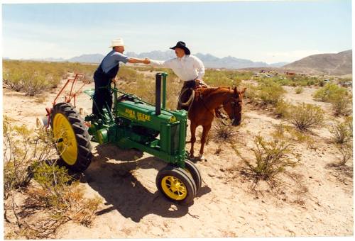 Two men interact in a desert landscape, one on a green tractor and the other on a horse, with mountains in the background.
