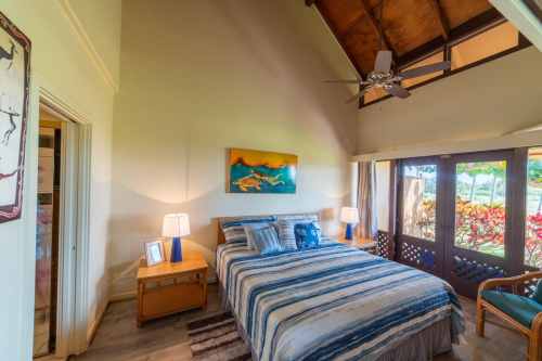 Cozy bedroom with a high ceiling, blue-striped bedding, wooden furniture, and large windows overlooking a garden.