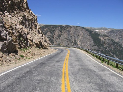 Winding mountain road with rocky cliffs on one side and green hills in the background under a clear blue sky.