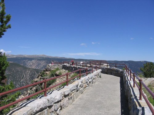 A scenic overlook with a stone pathway and railings, surrounded by mountains under a clear blue sky.