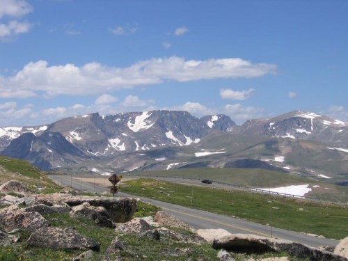 Scenic mountain landscape with snow-capped peaks, green meadows, and a winding road under a blue sky with clouds.