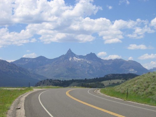 A winding road leads toward majestic mountains under a blue sky with fluffy clouds. Green hills frame the scene.