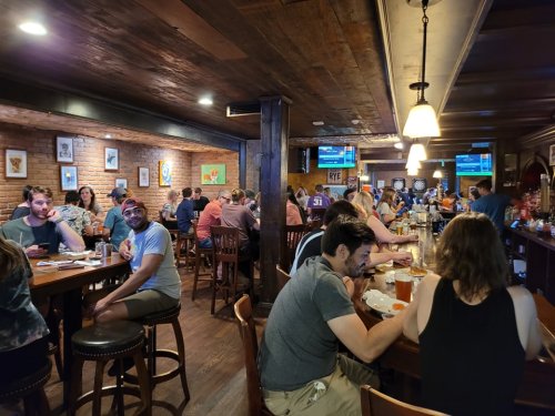 A lively bar scene with patrons enjoying drinks and food, seated at tables and the bar, with a warm, rustic atmosphere.