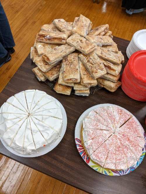 A table with a large pile of rectangular treats and two round cakes, one white and one red, sliced into pieces.