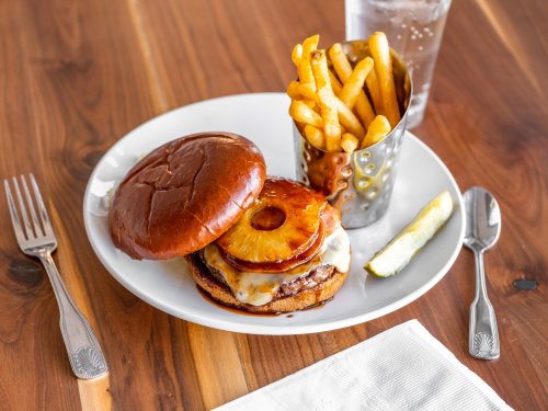 A burger with grilled pineapple and cheese, served with fries in a cup and a slice of lemon on a wooden table.