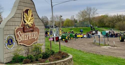 A welcome sign for Shepherd, with a gathering of people and tractors in the background on a cloudy day.
