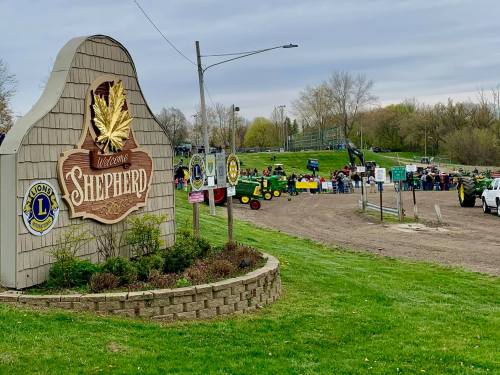 Welcome sign for Shepherd, with a crowd and tractors in the background on a grassy area under a cloudy sky.