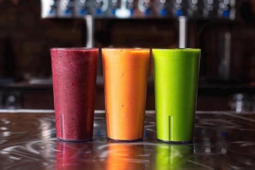 Three colorful smoothies in tall glasses: purple, orange, and green, on a shiny metal surface.