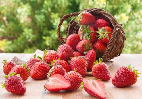 A basket of fresh strawberries spills onto a wooden surface, with some whole and one cut in half.