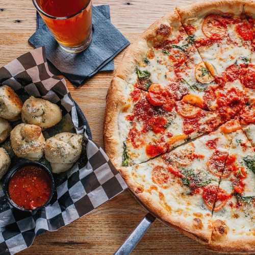 A table with a pizza topped with sauce and vegetables, alongside a basket of garlic knots and a drink.