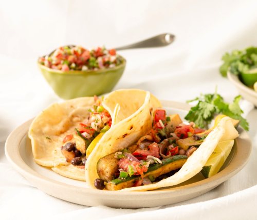 Three tacos filled with grilled vegetables and salsa, served on a plate with a side of fresh pico de gallo and lime.