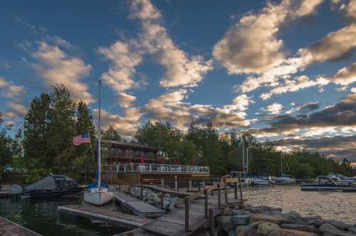 A scenic marina at sunset, featuring boats, a wooden dock, and a restaurant surrounded by trees and clouds.