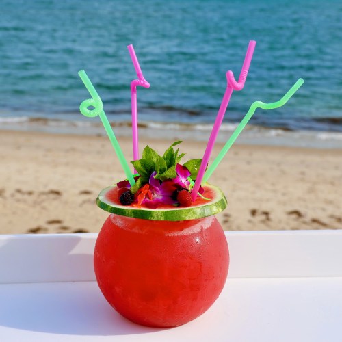A vibrant red cocktail in a watermelon bowl, garnished with flowers and berries, with colorful straws, by the beach.