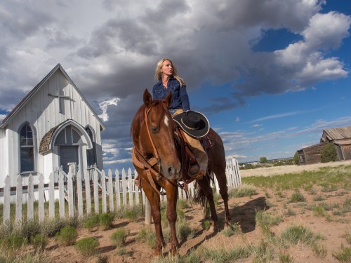 A woman on a horse stands near a white picket fence and a small church under a cloudy blue sky.