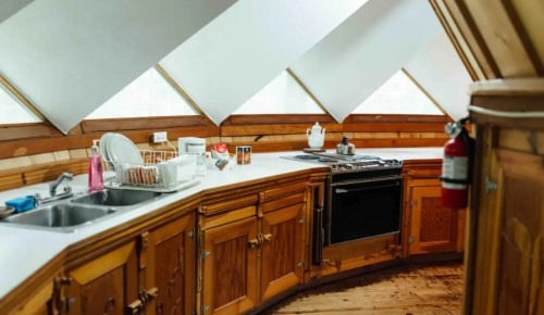 A cozy kitchen with wooden cabinets, a stove, sink, and large windows, featuring a unique angled design.