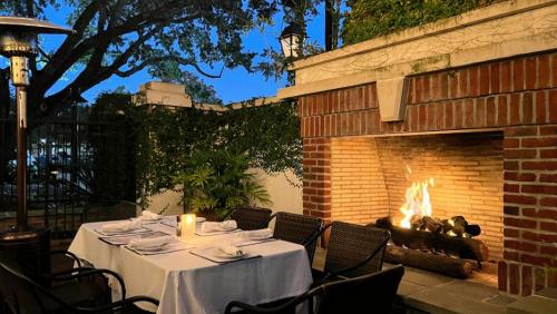 Cozy outdoor dining area with a table set for dinner beside a warm brick fireplace and lush greenery.