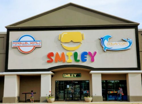 Colorful storefront featuring "SMILEY" sign, with logos for "Virtual Reality Center" and "Ocean Story." Kids playing outside.