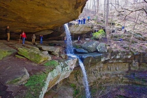 A waterfall cascades from a rocky overhang, with people exploring the area around it in a wooded setting.