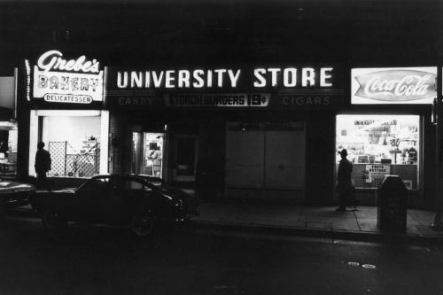 Black and white photo of a street at night featuring a bakery and a university store with neon signs.