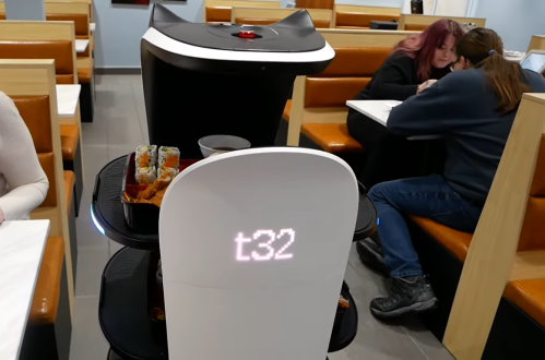 A robot server with a digital display delivers food in a restaurant, with diners seated at tables in the background.