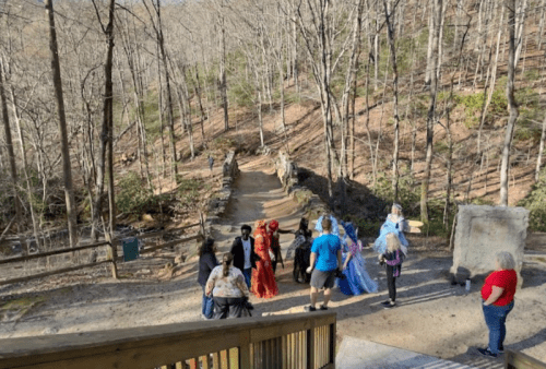 A group of people in costumes gathers on a forest path, surrounded by trees and a stone bridge in the background.