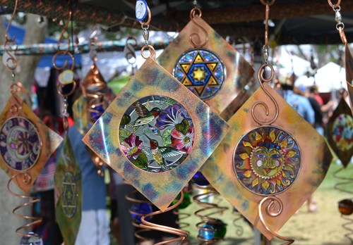 Colorful metal wind chimes with intricate designs hanging at a craft fair, surrounded by blurred festival attendees.