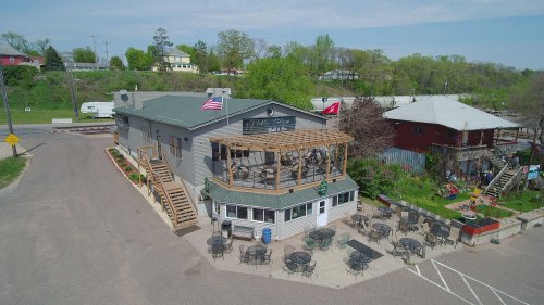 Aerial view of a restaurant with outdoor seating, surrounded by greenery and a few nearby buildings.