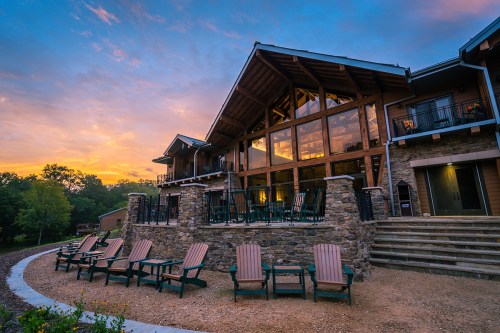 A rustic lodge with large windows and a stone patio, surrounded by chairs, set against a colorful sunset sky.