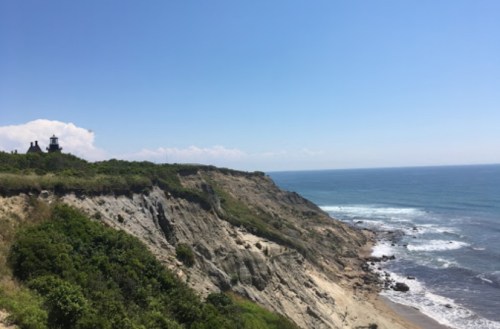 Coastal view featuring cliffs, greenery, and a lighthouse under a clear blue sky with ocean waves below.