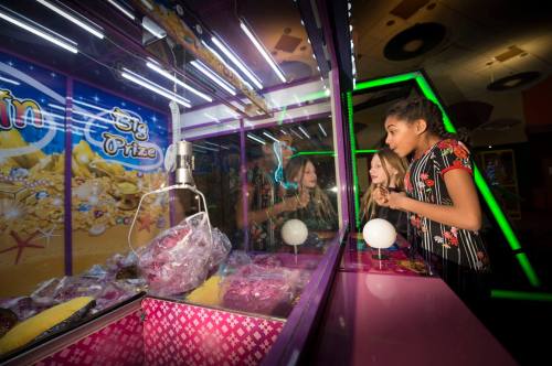 Two girls excitedly watch a claw machine filled with prizes in a colorful arcade setting.