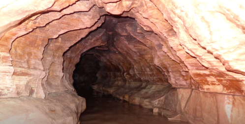 A narrow cave with smooth, layered rock walls and a small stream of water running through it.