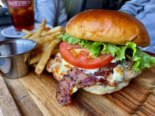 A close-up of a chicken sandwich with bacon, lettuce, and tomato, served with fries on a wooden board.