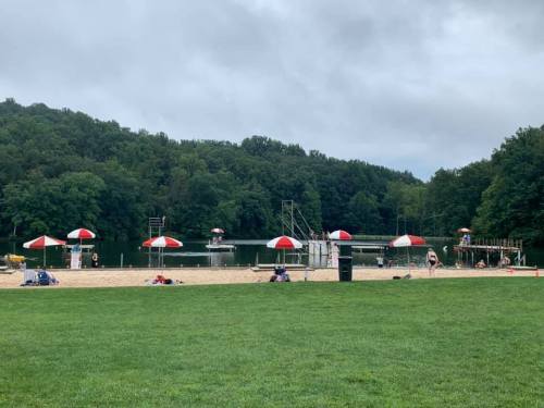 A sandy beach with red and white umbrellas, a dock, and people enjoying the water, surrounded by green trees under a cloudy sky.