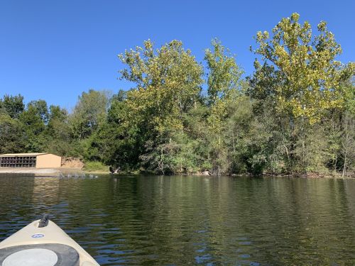 A view from a kayak on calm water, with trees and a building in the background under a clear blue sky.