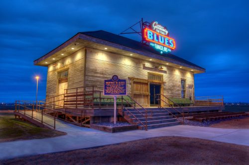 Historic building with a neon "Blues" sign, surrounded by a grassy area, under a twilight sky.