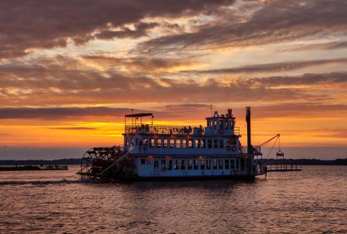 A paddleboat glides across the water at sunset, with vibrant orange and purple hues in the sky.
