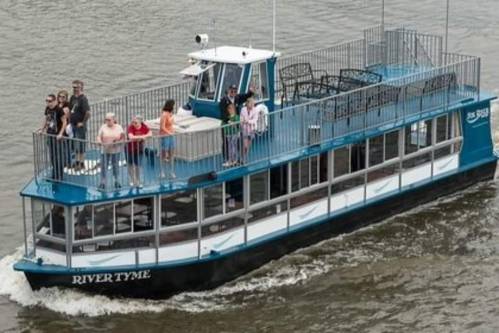 A blue and white riverboat named "RIVER TYME" with passengers enjoying the upper deck on a river.