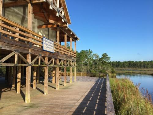 A wooden building on stilts overlooking a calm waterway, surrounded by trees and blue sky.