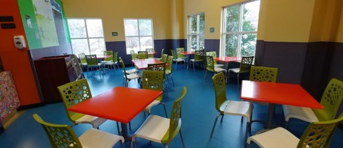 Brightly colored dining area with orange and green tables and chairs, featuring large windows and a vibrant atmosphere.
