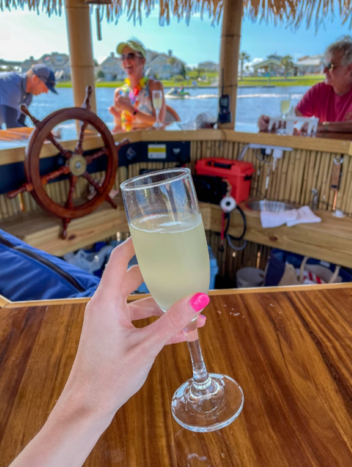 A hand holds a glass of drink in a tropical bar setting, with people and water in the background.