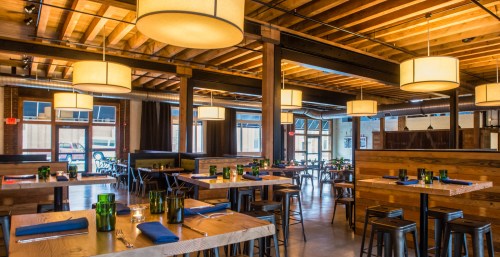 A spacious restaurant interior with wooden tables, green glassware, and pendant lights hanging from a wooden ceiling.