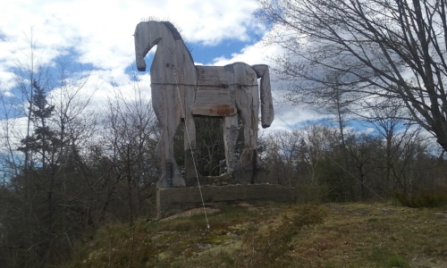 A large wooden sculpture of a horse stands on a grassy hill, surrounded by trees under a cloudy sky.