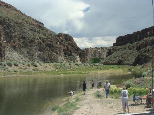 A scenic riverbank with people fishing and enjoying nature, surrounded by rocky cliffs and greenery under a cloudy sky.