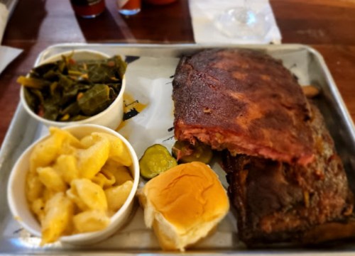 A tray with ribs, macaroni and cheese, collard greens, pickles, and a bun.